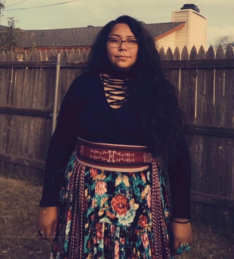 A person with long black hair stands in a fenced-in backyard. They are wearing a long floral skirt, tied with a red and white sash, and a black top.