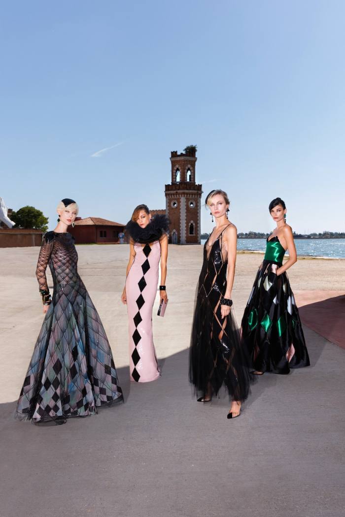 Models pose in full-length dresses incorporating harlequin patterns on a Venetian dock 