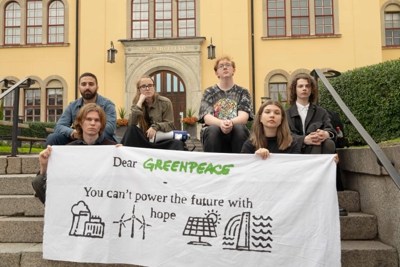 Six young people sit on stone steps outside a building, holding a banner that reads: 'Dear Greenpeace, You can't power the future with hope'