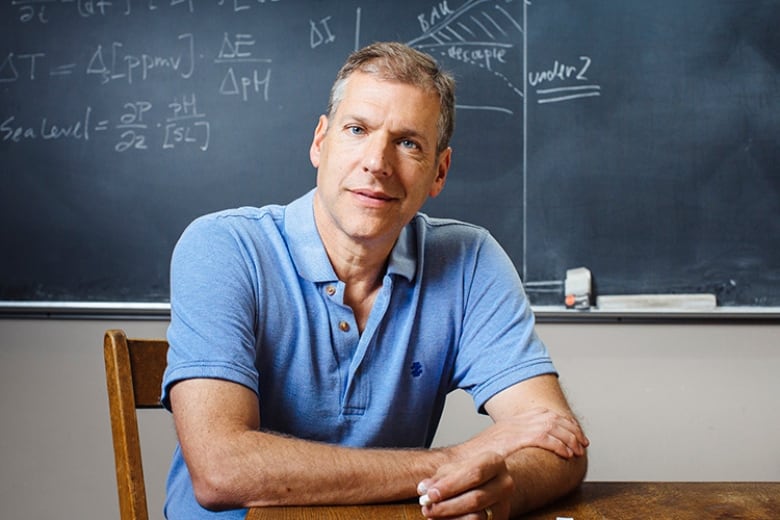 Portrait of a man in a blue polo shirt sitting in a classroom in front of a chalk board covered in math equations.