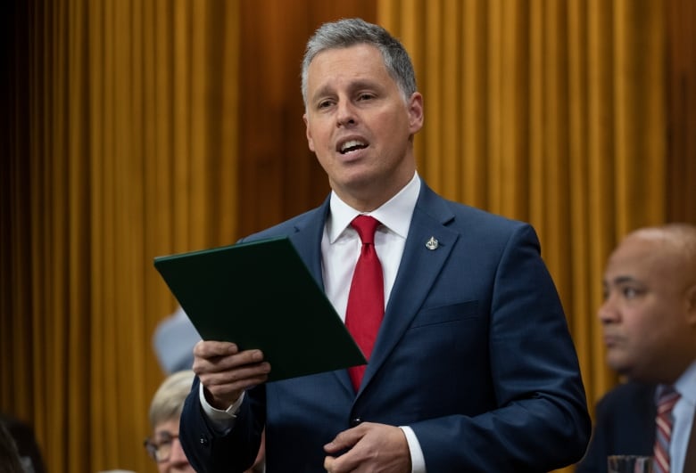 A man in a suit and red tie reads from a tablet in the House of Commons.