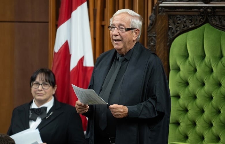 A man in a black robe, shirt and tie stands in the Speaker's chair in the House of Commons.