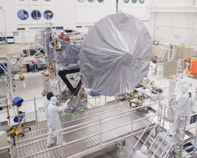 A huge antenna that's about twice as tall as the people who are attaching it to the Europa Clipper spacecraft is wrapped in white plastic in a clean room inside a large assembly facility. The people in the photo are dressed in white hazmat-looking suits.  