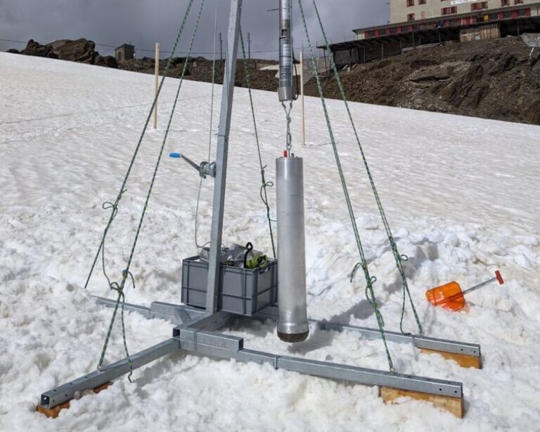 A small melting probe prototype is hung from a stand over a snowy surface with a building in the distant background.