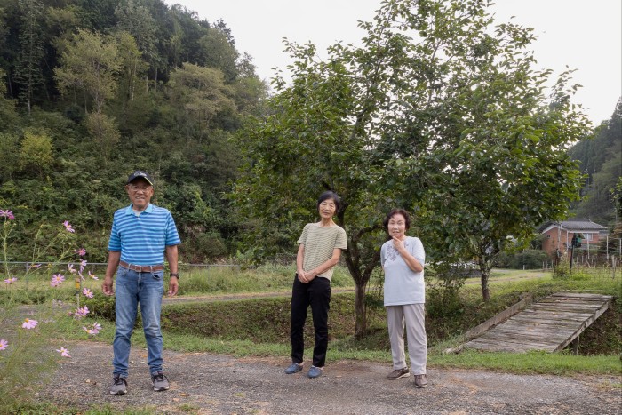 A man and two women, probably in their seventies, stand in a rural setting with a small wooden bridge behind them