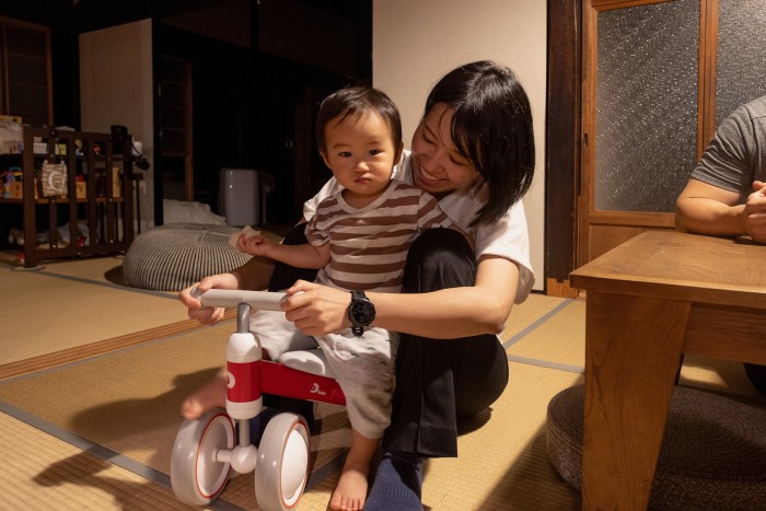 A woman sits on the floor, her arms around a little boy who sits on a wooden trike