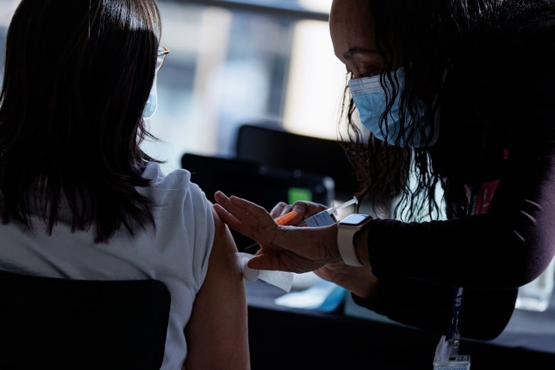 A nurse wearing a blue medical face mask pushes a needle into a young woman's upper arm.