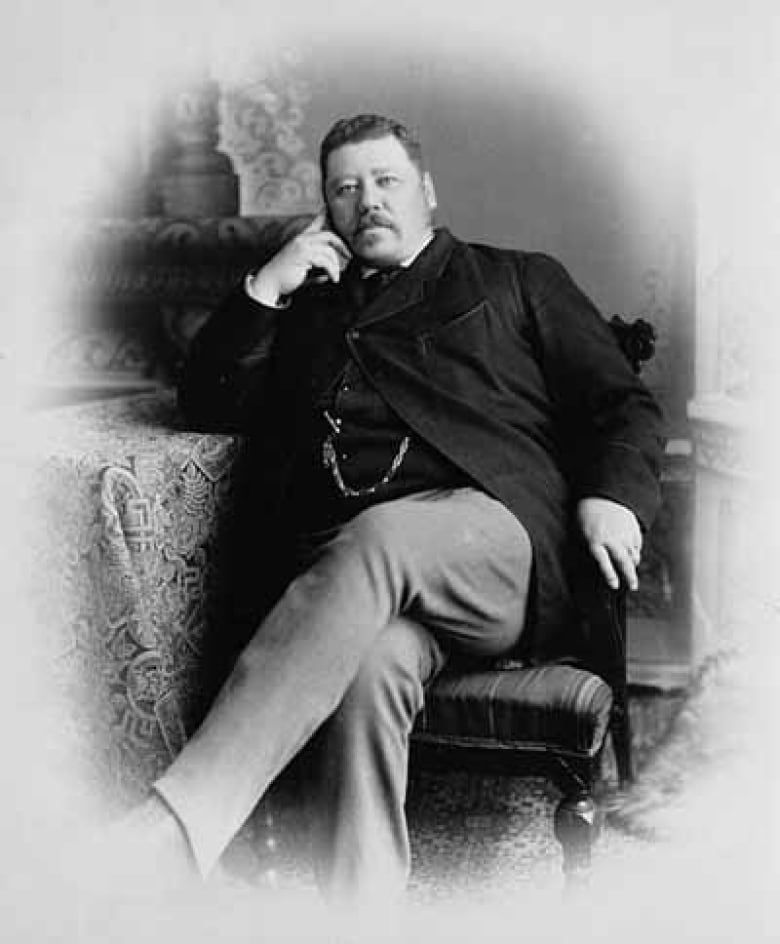 Black and white photo of a man sitting in a chair, his head slightly tilted and resting on his right hand. He's got a black suit jacket and vest, with a watch chain seen looped from the vest.