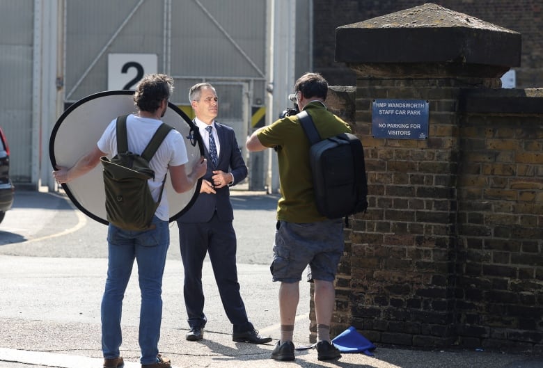 A male TV reporter in a suit stands in front of Wandsworth prison, London, U.K., facing a lighting crew worker and a cameraman 