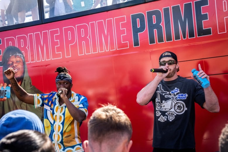 Two men stand in front of a red bus talking into microphones and holding cans.