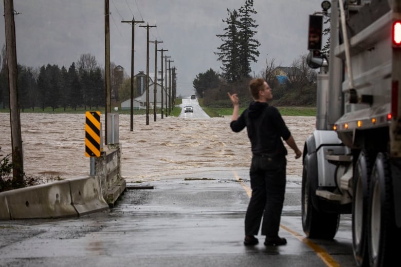 A man stands next to a truck and points at a road that is completely submerged by flood waters.