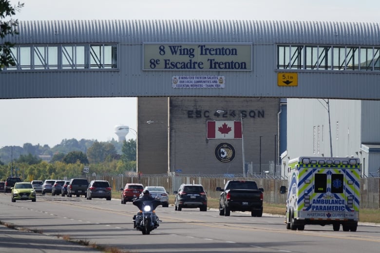 Traffic, including a motorcycle and ambulance, can be seen driving in both directions under a large, covered, pedestrian overpass with the words 8 Wing Trenton on the side in big letters.