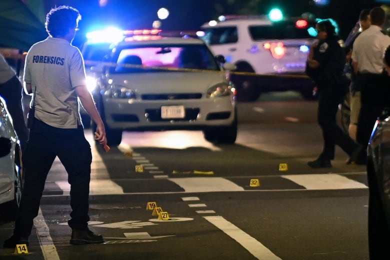 A night street scene with a man wearing a shirt that reads "forensics" and police cars.