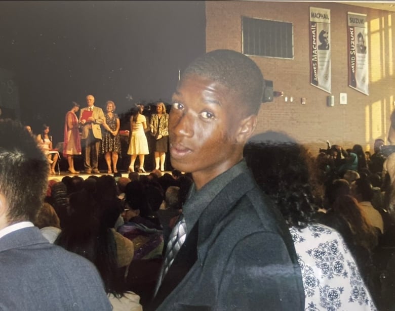 A teenager in a suit looks back at the camera while a group of elementary school students stand on stage.