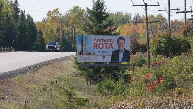 A sign on the side of the road says "Anthony Rota, MP." A truck is driving in the background.