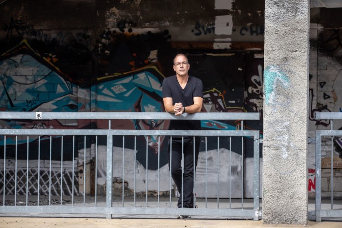 A man stands leaning against a metal railing; behind him is a concrete wall covered with graffiti