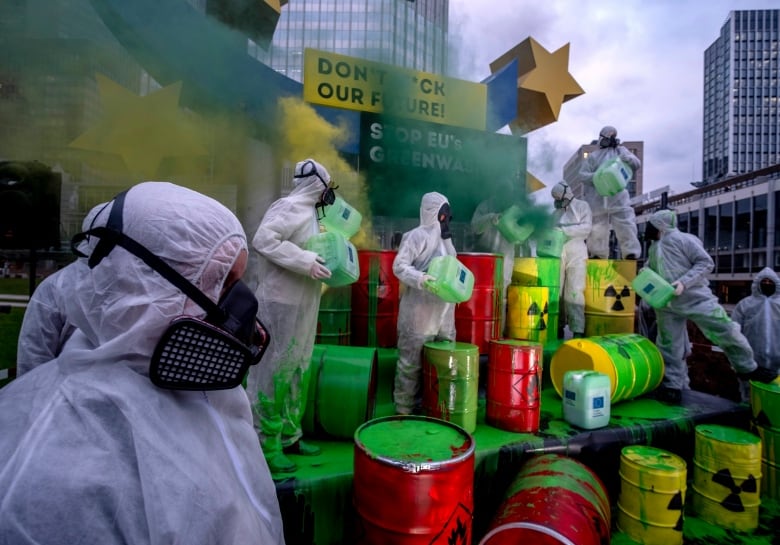 People wear hazmat suits and masks, and carry vats with toxic symbols on them covered in bright green paint, meant to look like toxic waste.  Signs behind them read, 'Don't f*ck our future' and 'Stop EU's Greenwashing.'