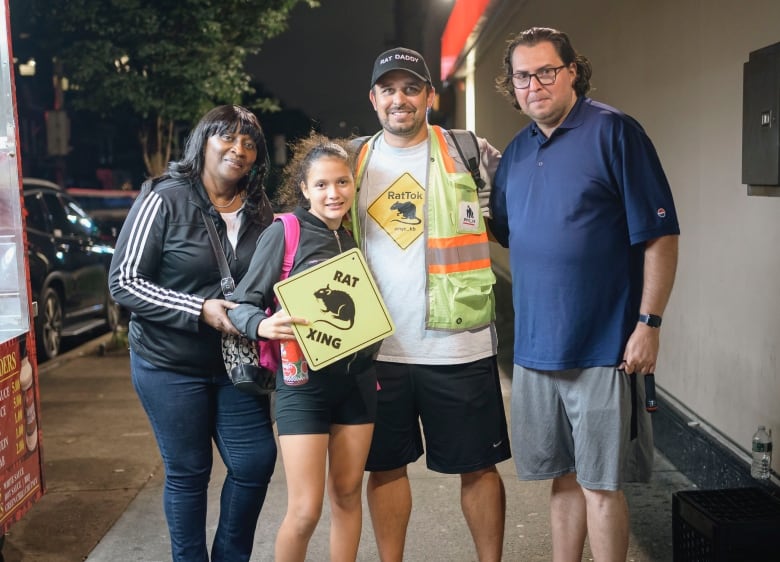 A man in a safety vest sports a T-shirt that with a rat silhouette in a yellow diamond that reads "RatTok." He's standing outside a food truck, posing with a man, a woman and a young girl.