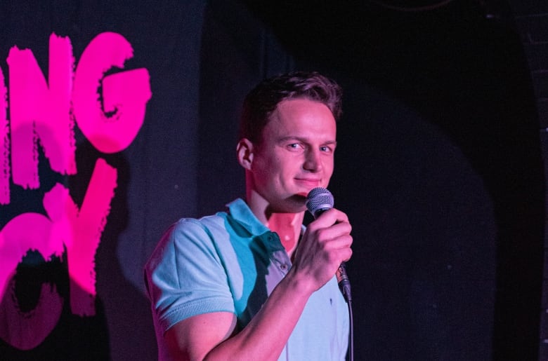 A man wearing a blue polo shirt smiles while holding a microphone on stage. 