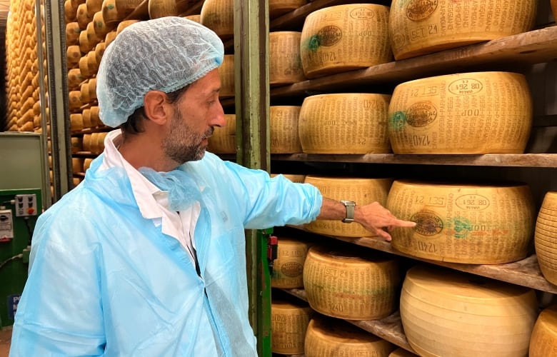 A bearded man wearing a light blue uniform points at a wheel of cheese.