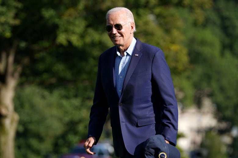 A man with white hair, wearing sunglasses and dressed in a navy blue suit jacket and blue shirt, walks in the outdoors, smiling.