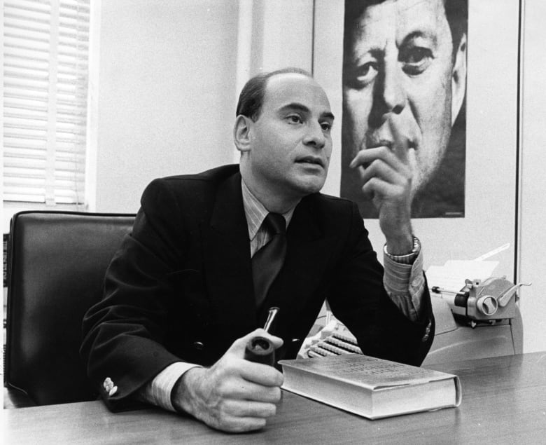 A man in a suit and tie is shown sitting at a desk in a black and white photograph.