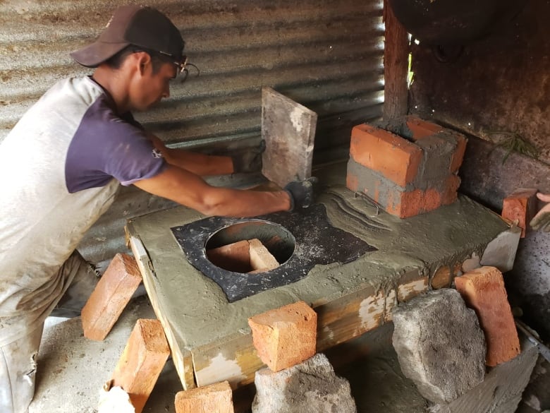 A trainee is spreading cement across a cement and brick base for a stove.