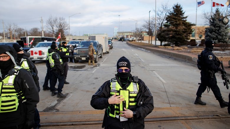 Police officers in bright neon vests stand in front of a row of pickup trucks with Canadian flags on them in front of a border crossing.