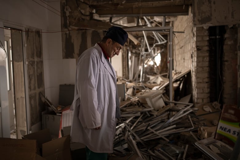 A man in a white coat stands with his head bowed in a destroyed building