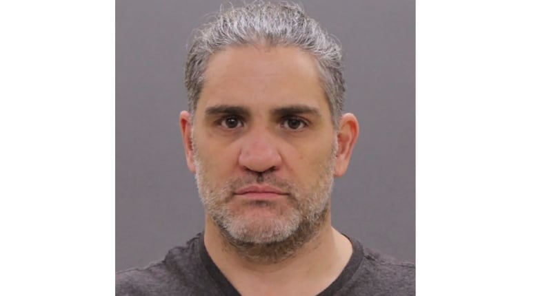 A police mug shot of a grey haired man with facial hair, wearing a grey tee shirt.