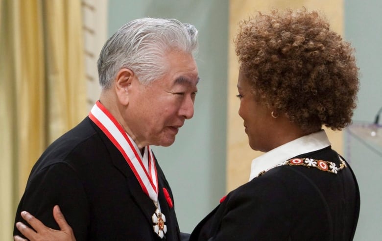 A woman with curly brown hair embraces an older Japanese man wearing an Order of Canada medal. 