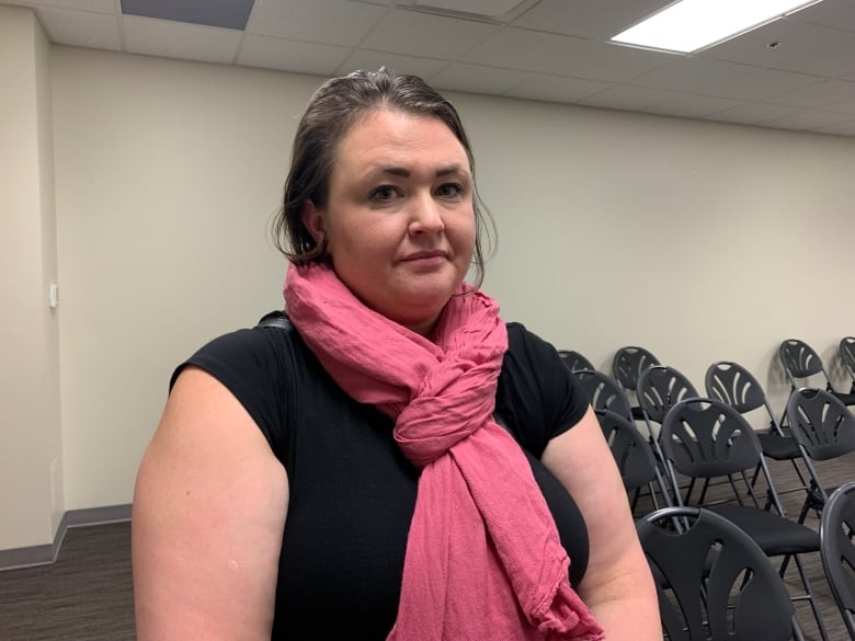 A woman with brown hair, dressed in a black shirt and pink scarf, looks into the camera.