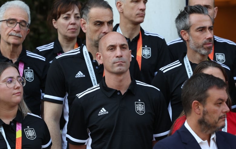 A man in a black polo stands with his hands behind his back.