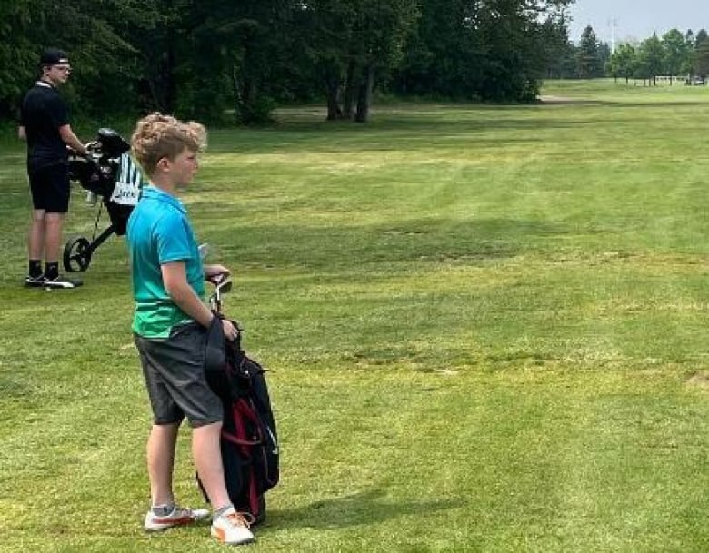 A boy stands on a golf course next to a golf bag. 