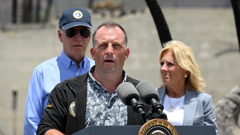 A person speaks at a lectern outside as two people behind the speaker watch on.