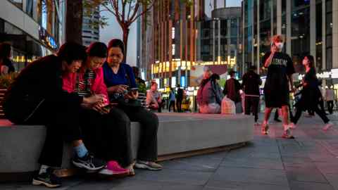 Women sit together as they watch a video on a mobile phone in a busy retail shopping area n Beijing, China