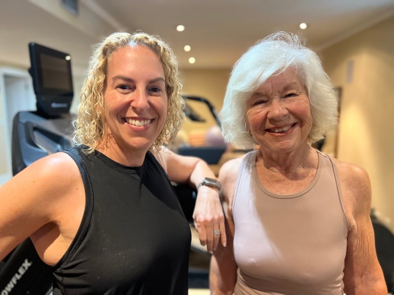 Two women, one with blond, curly hair, the other with a white bob, smile at the camera while wearing workout clothing and standing in a gym. 