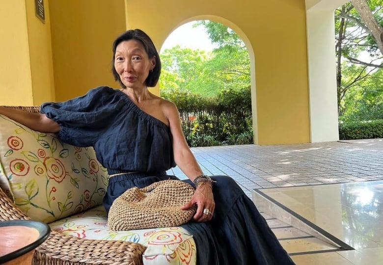 A woman with a chin-length bob wears a blue one-shoulder dress and lounges in a wicker seat in a yellow room. 