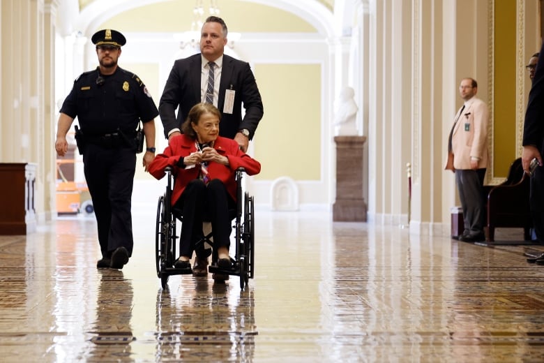 Woman in wheelchair being wheeled down a hall by a man who is accompanied by security. 