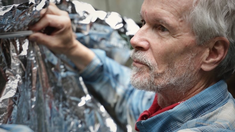 An grey-haired man in a blue plaid shirt attaches a foil blanket to a window.