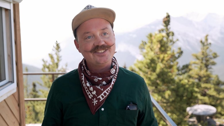 A man in a dark green shirt and a colorful scarf is seen smiling at the camera.