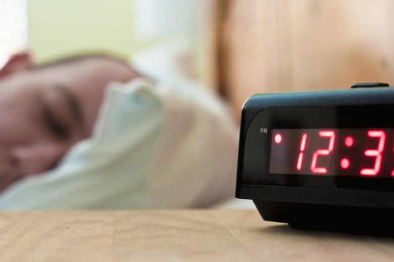 A man's head is seen resting on a pillow while a bedside alarm clock reads 12:30 p.m.