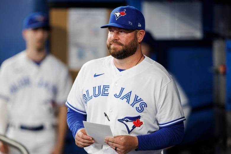 A bearded man wearing a white baseball uniform and blue baseball hat holds a sheet of paper.