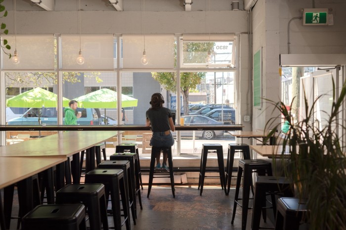 Blond-wood tables and stools inside Faculty Brewing Co, with a woman sitting at a counter at the floor-to-ceiling windows