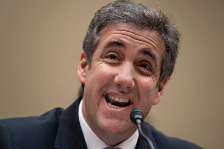 A man in a suit and tie is shown speaking into a microphone while seated at a table.