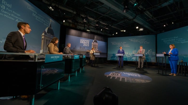 Three people are seated at desks while four others stand at podiums in a TV studio.