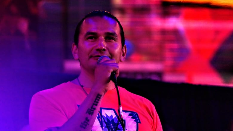 A man wearing an orange T-shirt is standing on a stage and speaking into a microphone.