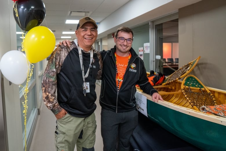 Two men stand together beside a canoe in a hallway. They are smiling.