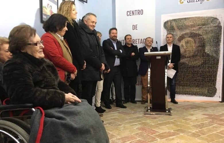 A group of people stand next to a podium and large print of a 19-century Spanish painting of Christ. An elderly woman in a wheelchair smiles in the foreground. 