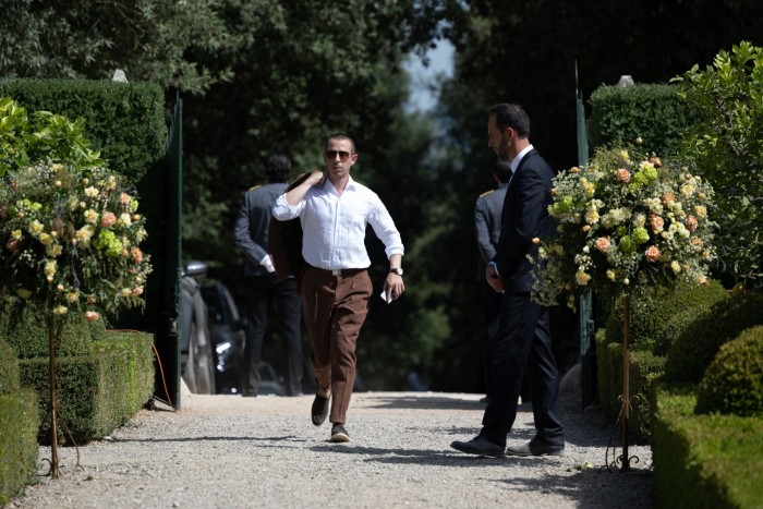 A man in white shirt and brown trousers walks through a garden, his jacket slung over his shoulder